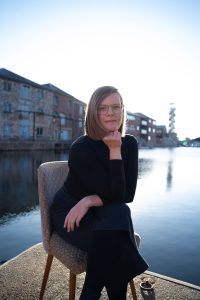 A woman with light skin, shoulder length straight dark blonde hair and round glasses is sat on a patterned dining chair by the water on a quayside. She has a small smile, one hand folded under her chin and other draped across her lap. The background is out of focus but across the water there is a row of large red brick buildings.
